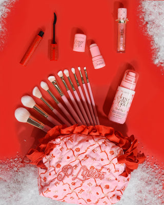 Pink Christmas P.Louise makeup bag with red festive design on it with makeup products coming out the top including makeup brushes, lip oil, mascara and more on a red background with snow