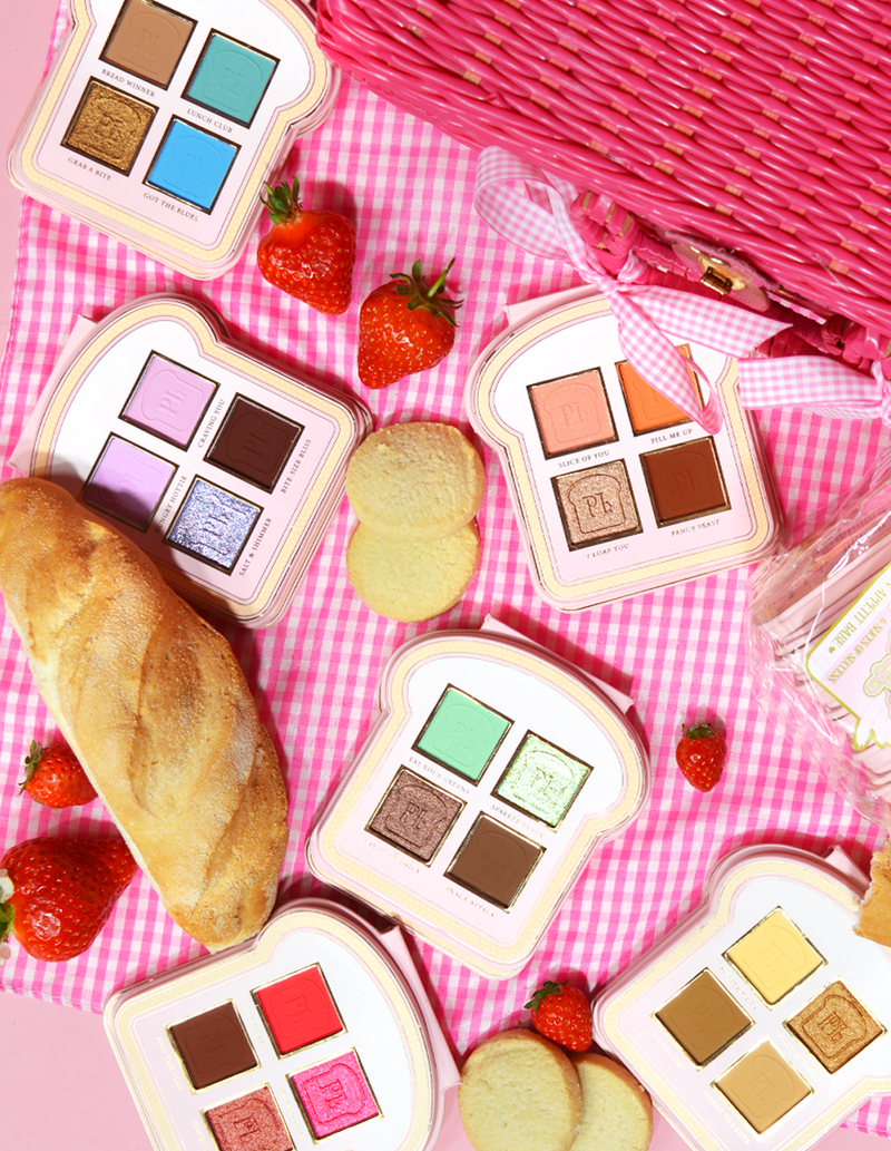 Un ensemble de mini palettes de fards à paupières de style sandwich avec différentes nuances de couleurs de maquillage affichées sur une surface vichy rose, accompagnées de fraises et d'un morceau de pain.
