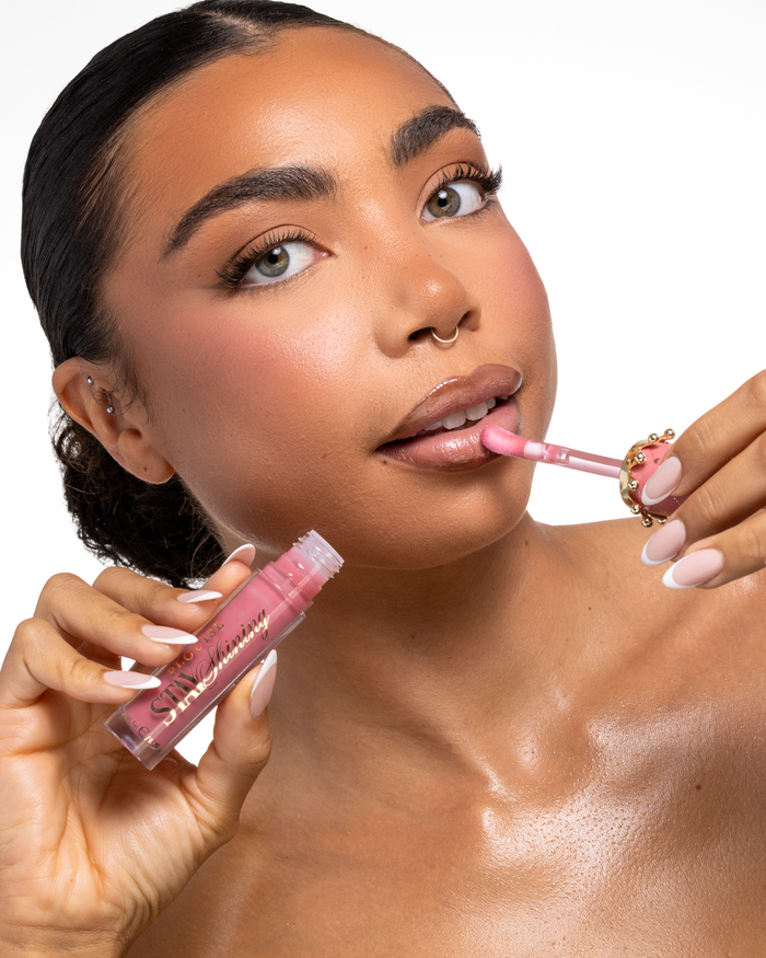 Een close-up van een vrouw die een roze P.Louise Stay Shining Lip Oil vloeibaar lipproduct aanbrengt met een doe-voetapplicator terwijl ze de tube in haar andere hand houdt, set tegen een schone witte achtergrond.