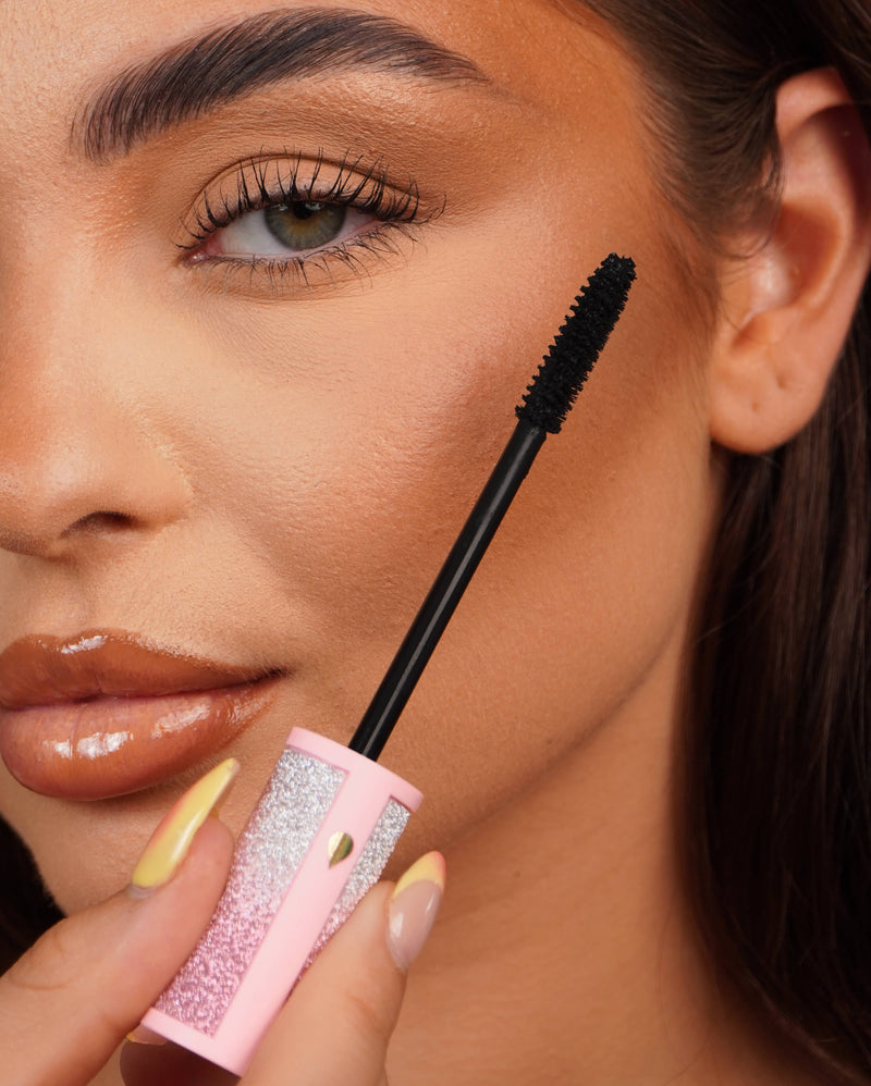 Gros plan d'une baguette de mascara P.Louise Cry Baby tenue près de l'œil d'une personne avec un tube de mascara rose pailleté.