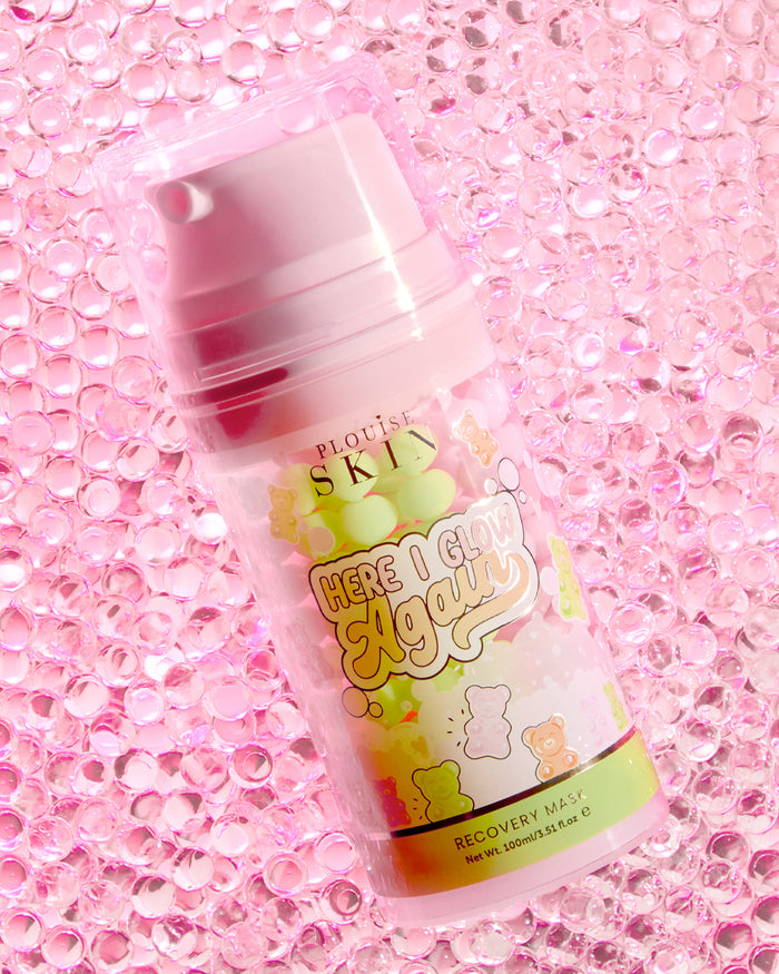 Een fles P.Louise Here I Glow Again Recovery Mask in de geur van snoepberen met een roze en groen label, met illustraties van snoepberen, geplaatst op een roze glitterachtergrond.