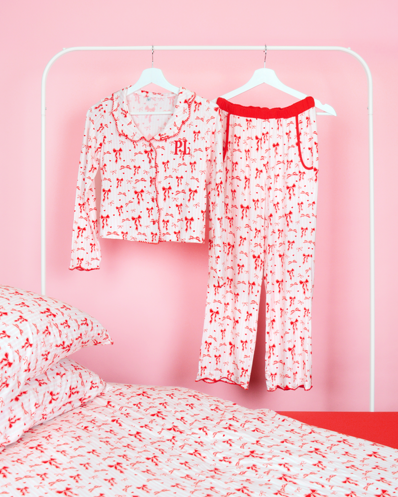 P.Louise kerstpyjamabroek met strik voor meisjes met een all-over rood strikontwerp tegen roze en witte strepen die aan kleerhangers hangen in een roze slaapkamer met bijpassend beddengoed