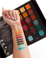 An open halloween eyeshadow palette displayed on a white background, with a person's forearm and hand resting beside it. The palette contains multiple square eyeshadow pans in warm oranges, reds, browns, blues, and greens, and the hand shows swatches of several of the shades applied across the forearm.
