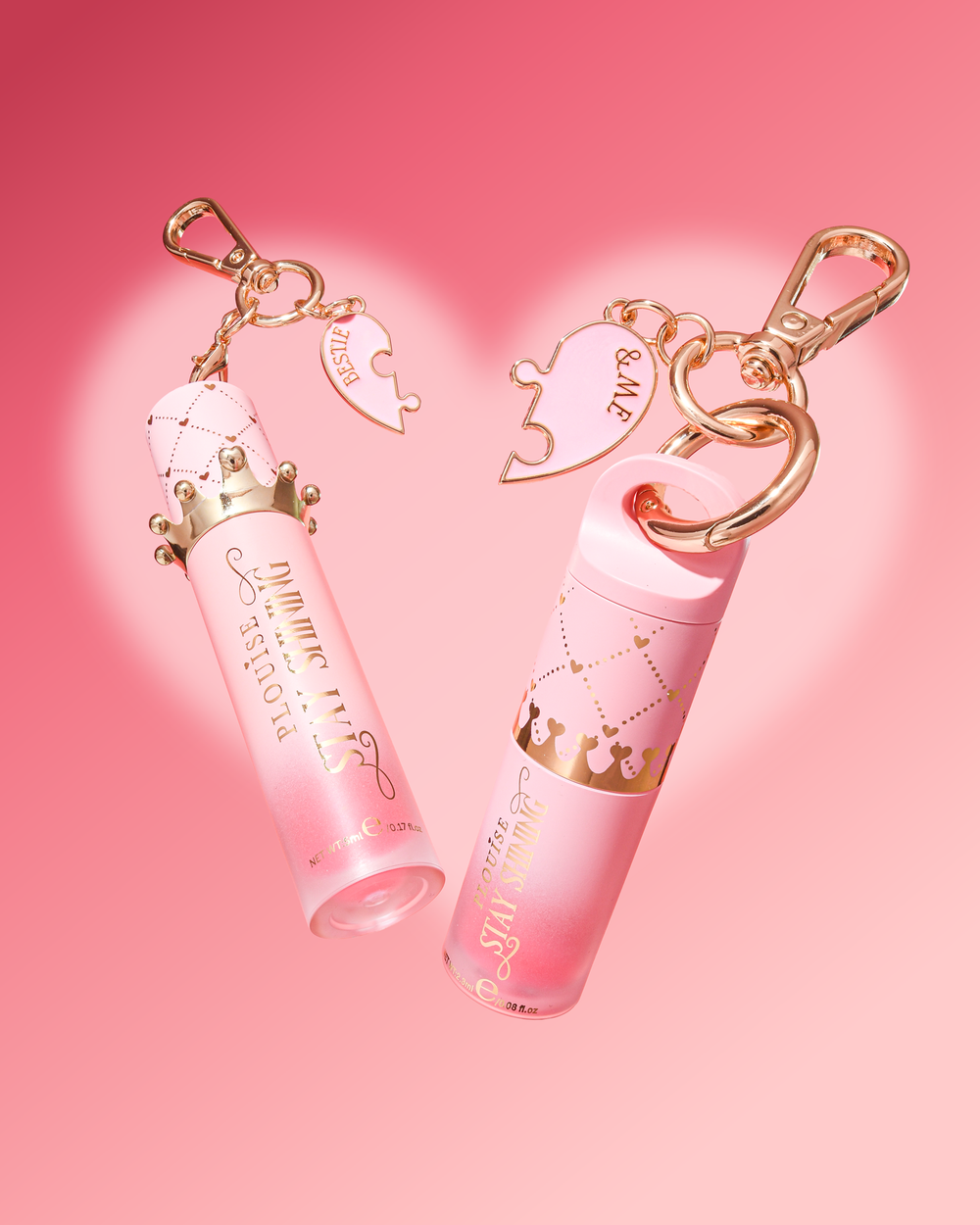 Two pink lip gloss tubes with gold-tone keychain clips and heart-shaped charms, displayed on a soft pink background with a faint heart outline.