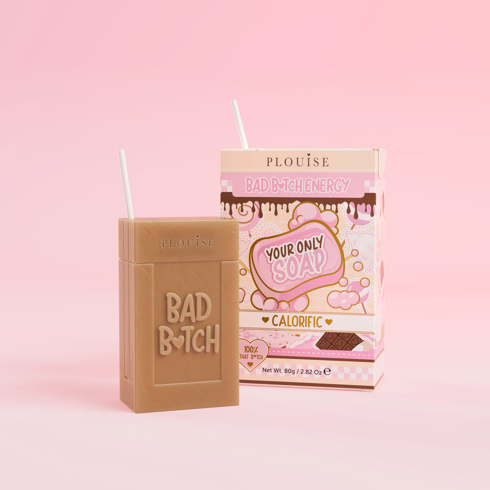 Un pain de savon marron sur lequel est gravé « Bad Bitch », accompagné d'un emballage de boîte de jus coloré portant l'inscription « Calorific », présenté sur fond rose.