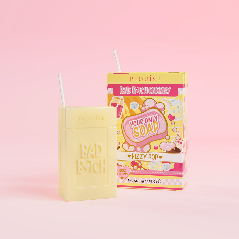 Un pain de savon jaune sur lequel est gravé « Bad Bitch », accompagné d'un emballage de boîte de jus coloré portant l'inscription « Fizzy Pop », présenté sur un fond rose.