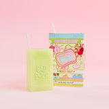 Un pain de savon vert sur lequel est gravé « Bad Bitch », accompagné d'un emballage de boîte de jus coloré portant l'inscription « Mixed Fruit », présenté sur un fond rose.