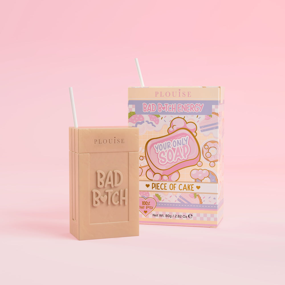 Un pain de savon beige sur lequel est gravé « Bad Bitch », accompagné d'un emballage de boîte de jus coloré portant l'inscription « Piece of Cake », présenté sur un fond rose.