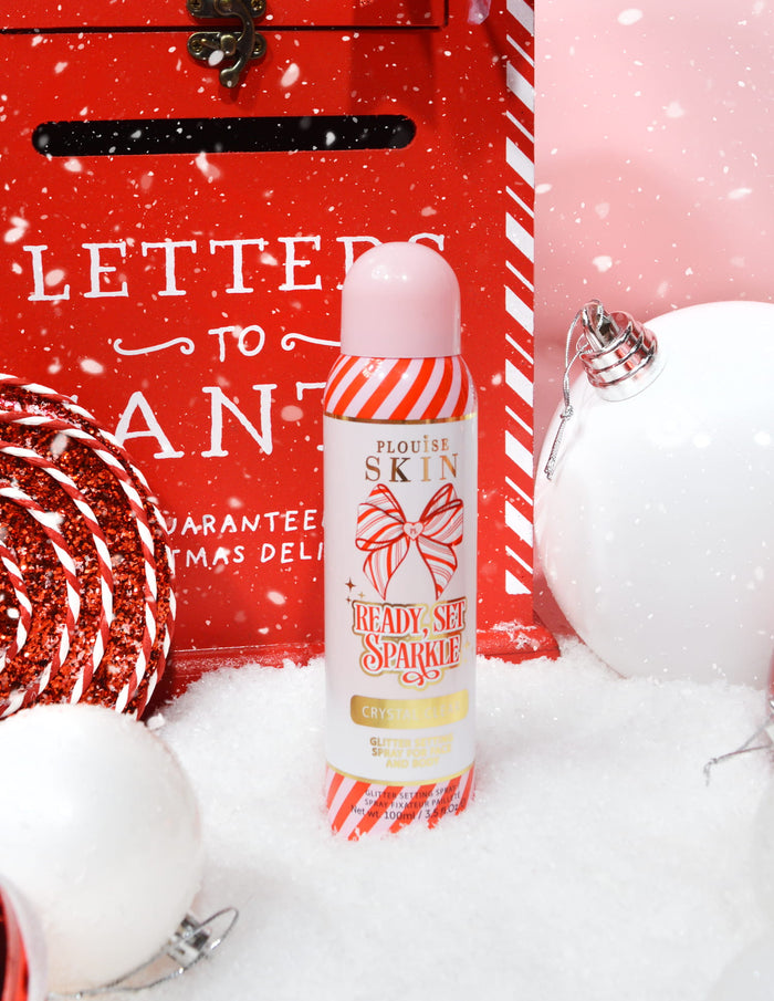 Een fles P. Louise Ready Set Sparkle Christmas Glitter Setting Spray omringd door feestelijke versieringen zoals sneeuw, kerstballen en een kerstbrievenbus