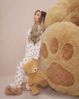 Vrouw in teddypyjama die een teddybeer vasthoudt naast een grote pluchen beerpoot op een beige achtergrond