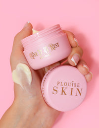 Une personne tenant un pot rose de crème pour les yeux P.Louise Give It A Rest sans le couvercle, affichant le nom du produit et la marque sur le pot.