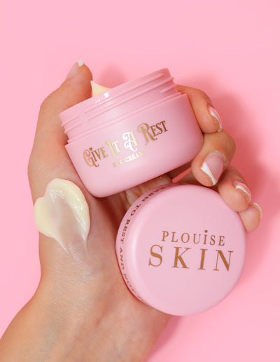 Une personne tenant un pot rose de crème pour les yeux P.Louise Give It A Rest sans le couvercle, affichant le nom du produit et la marque sur le pot.